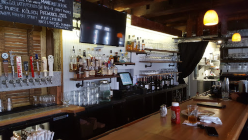 A rustic bar with a wooden counter, shelves of liquor, and a TV, featuring glasses and condiments on the counter.