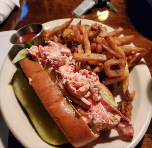 A lobster roll with lettuce on a bun, served with a side of curly fries and a small cup of dipping sauce.