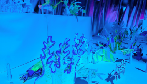 A vibrant underwater scene made of paper, featuring colorful fish and coral in shades of blue and purple light.