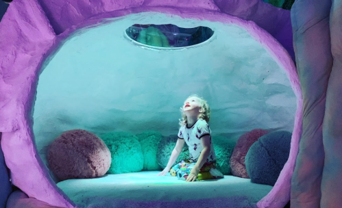 A child sits inside a colorful, cave-like structure, looking up with a joyful expression. Soft cushions surround them.