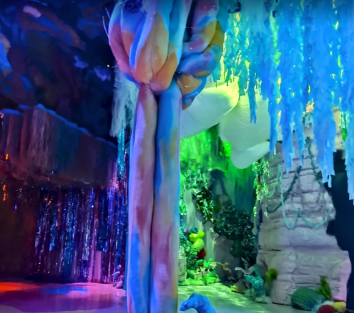 Colorful, abstract indoor landscape with vibrant trees and hanging decorations, illuminated by various lights.
