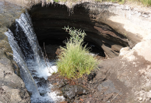 A small waterfall flows into a dark, circular hole in the ground, surrounded by grass and rocky terrain.