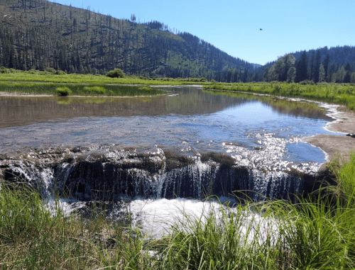 A serene landscape featuring a small waterfall flowing into a calm river, surrounded by lush greenery and mountains.