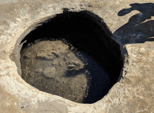 A deep, circular hole in the ground filled with murky water and debris, with a shadow of a person nearby.
