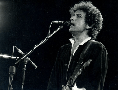A musician with curly hair sings into a microphone while playing guitar on stage, illuminated by stage lights.