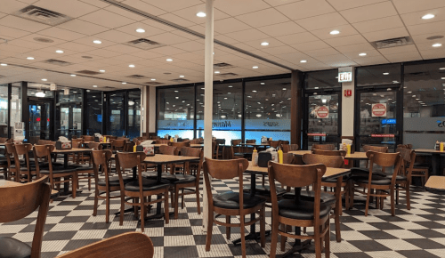 Empty diner interior with wooden chairs and tables, checkered floor, and large windows. Brightly lit and spacious.