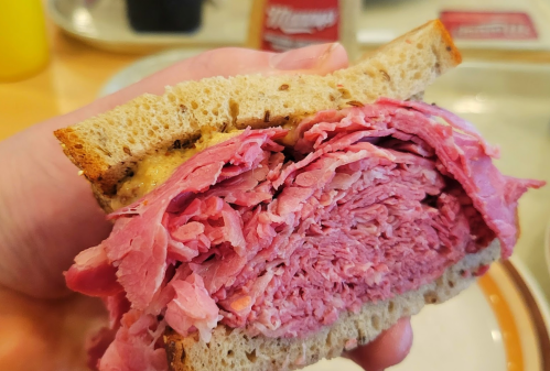 A hand holding a thick sandwich filled with layers of pink deli meat and mustard on whole grain bread.