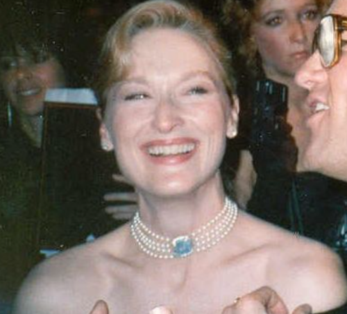 A smiling woman in a pearl necklace, surrounded by people at an event, exuding joy and elegance.