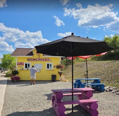 A colorful yellow food stand with an umbrella and picnic tables, set against a blue sky with fluffy clouds.