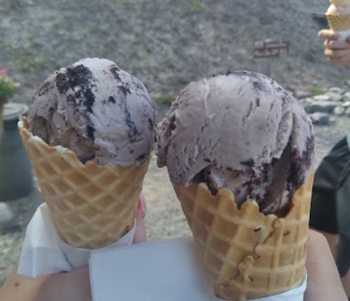 Two ice cream cones with chocolate and cookies and cream flavors, held in a hand against a rocky background.