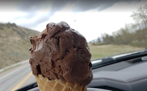 A close-up of a chocolate ice cream cone held inside a car, with a scenic landscape visible outside the window.