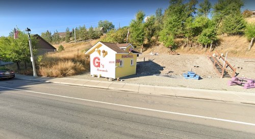 A small yellow building with "G's" signage, set against a hillside, near a road with trees in the background.
