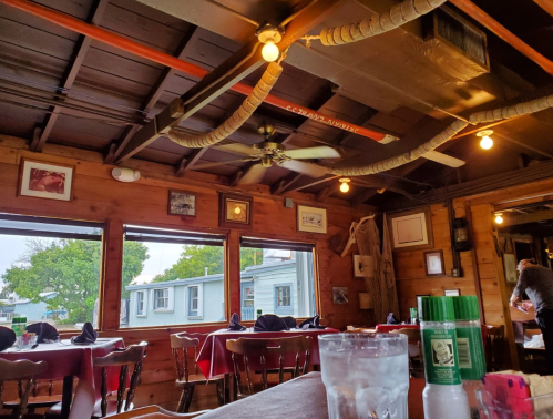 Cozy restaurant interior with wooden walls, ceiling fans, and large windows showing an outdoor view.