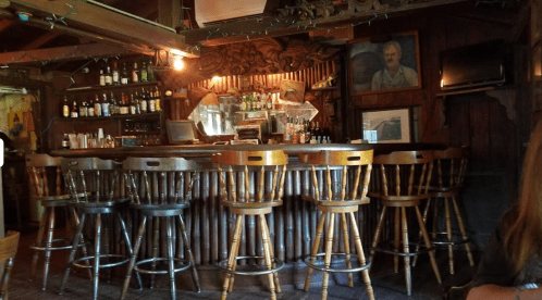 A rustic bar with wooden stools, shelves stocked with bottles, and a vintage portrait on the wall.