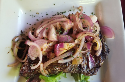 Grilled steak topped with sliced onions and herbs, served on a white plate with a bed of lettuce.