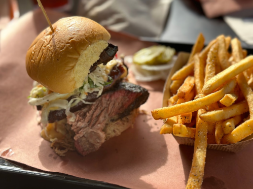 A close-up of a beef sandwich with coleslaw on a bun, served with a side of crispy fries and a pickle.