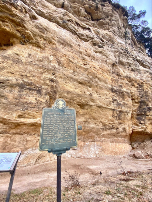 A historical marker stands in front of a large, rocky cliff face, surrounded by sparse vegetation.