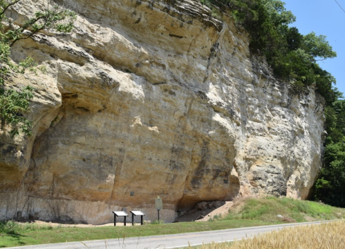 A large rock formation beside a road, with informational signs and greenery in the background.
