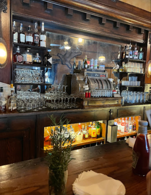 A vintage bar with shelves of liquor bottles, glassware, and a classic cash register, set against a wooden backdrop.