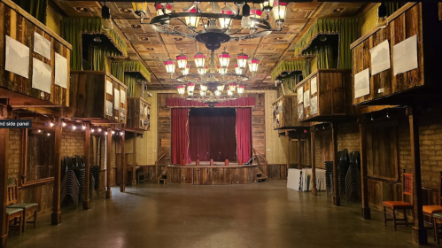Interior of a vintage theater with wooden accents, stage curtains, and balcony seating. Dimly lit with hanging lights.