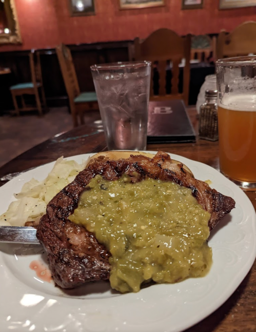 A plate with a grilled steak topped with green sauce, served with a side of cabbage, a glass of water, and a beer.