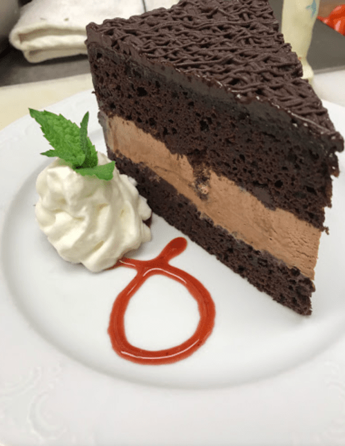 A slice of chocolate cake with a creamy filling, topped with whipped cream and a mint leaf, drizzled with sauce.