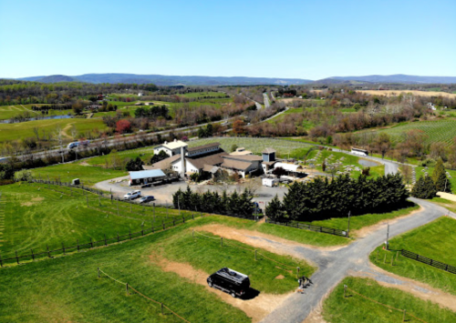 Aerial view of a farm with fields, a building complex, and surrounding greenery under a clear blue sky.