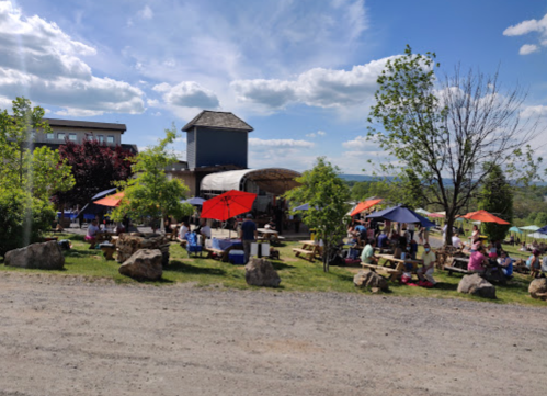 A lively outdoor scene with people sitting at picnic tables under colorful umbrellas, surrounded by trees and a blue sky.