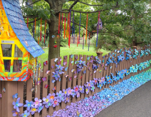 Colorful fence adorned with flowers and butterflies, with a whimsical house and playful decorations hanging from a tree.