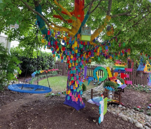 A colorful tree adorned with vibrant decorations and a swing, surrounded by greenery and playful art elements.