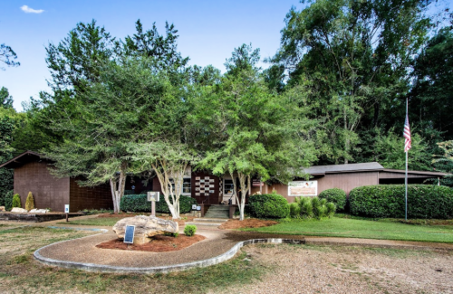 A rustic building surrounded by trees and shrubs, featuring a flagpole and a gravel pathway in front.