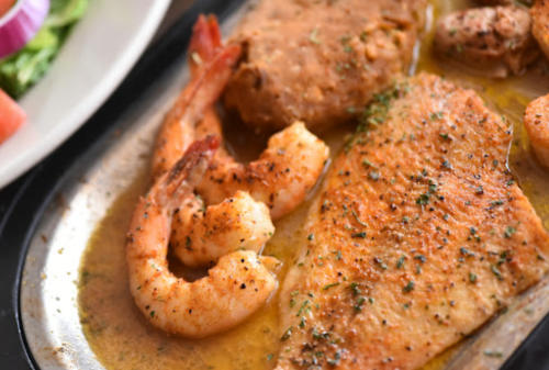 A close-up of a sizzling seafood platter featuring shrimp and fish, garnished with herbs in a savory sauce.