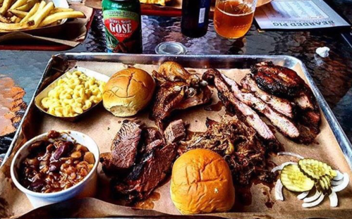 A metal tray filled with various BBQ meats, macaroni and cheese, baked beans, and two buns, with drinks in the background.