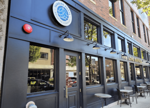 Exterior of Tangled Root Brewing Company, featuring large windows and a sign, with outdoor seating visible.