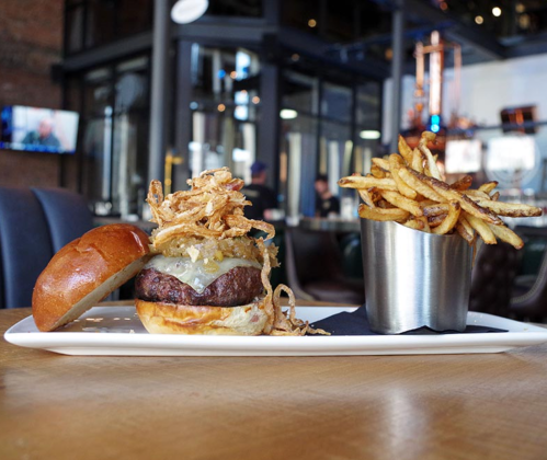 A gourmet burger topped with crispy onions and cheese, served with a side of golden fries in a metal cup.