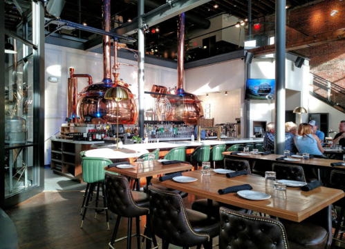 A modern restaurant interior featuring copper brewing tanks, wooden tables, and patrons enjoying their meals.