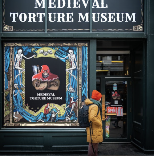 A person in a yellow coat and orange hat stands in front of the Medieval Torture Museum, featuring a colorful sign.
