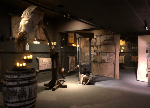 A dimly lit room featuring medieval torture devices, eerie decor, and flickering candles, creating a haunting atmosphere.