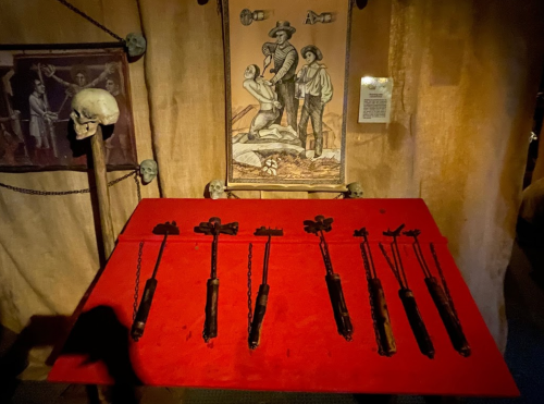 A display of various historical torture devices on a red table, with skulls and a framed illustration in the background.