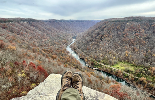 A pair of hiking boots on a rocky ledge overlooking a winding river and autumn-colored mountains.