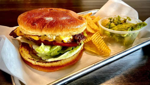A juicy burger with lettuce, tomato, and cheese, served with wavy potato chips and a side of guacamole.