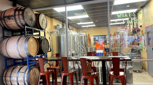Interior of a brewery featuring barrels, brewing equipment, and a seating area with metal chairs.
