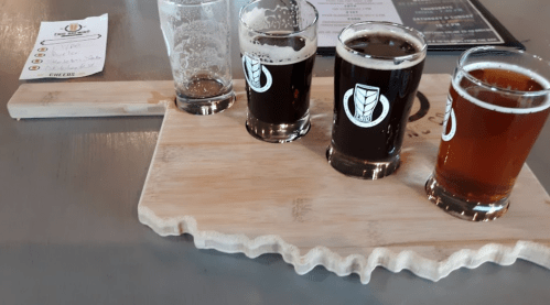 Three craft beer samples on a wooden paddle shaped like Oklahoma, with a glass and a menu in the background.