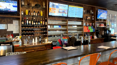 A bar with wooden shelves stocked with drinks, two TVs showing sports, and a staff member cleaning behind the counter.