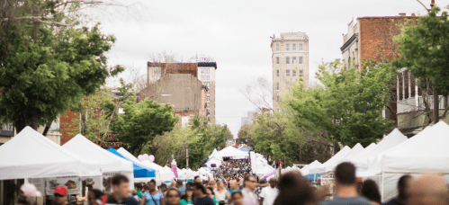 A bustling street fair with crowds and vendor tents lining the road, surrounded by trees and buildings.