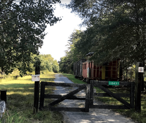 A gravel path leads to a wooden gate, with a vintage train car visible among trees in the background.