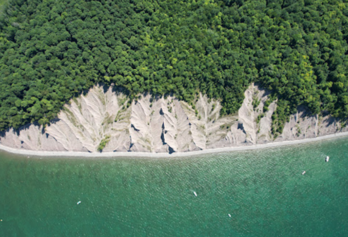 Aerial view of a coastline with layered sandy cliffs and lush green forest above, alongside calm turquoise waters.