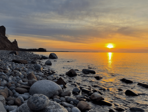 A serene sunset over a calm lake, with smooth stones lining the shore and soft clouds in the sky.