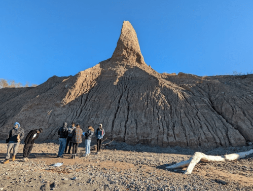 A group of people stands on a rocky beach in front of a tall, pointed rock formation under a clear blue sky.
