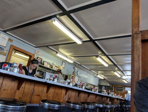 A diner interior with a counter, staff serving customers, and a casual atmosphere. Bright lights and wooden decor.